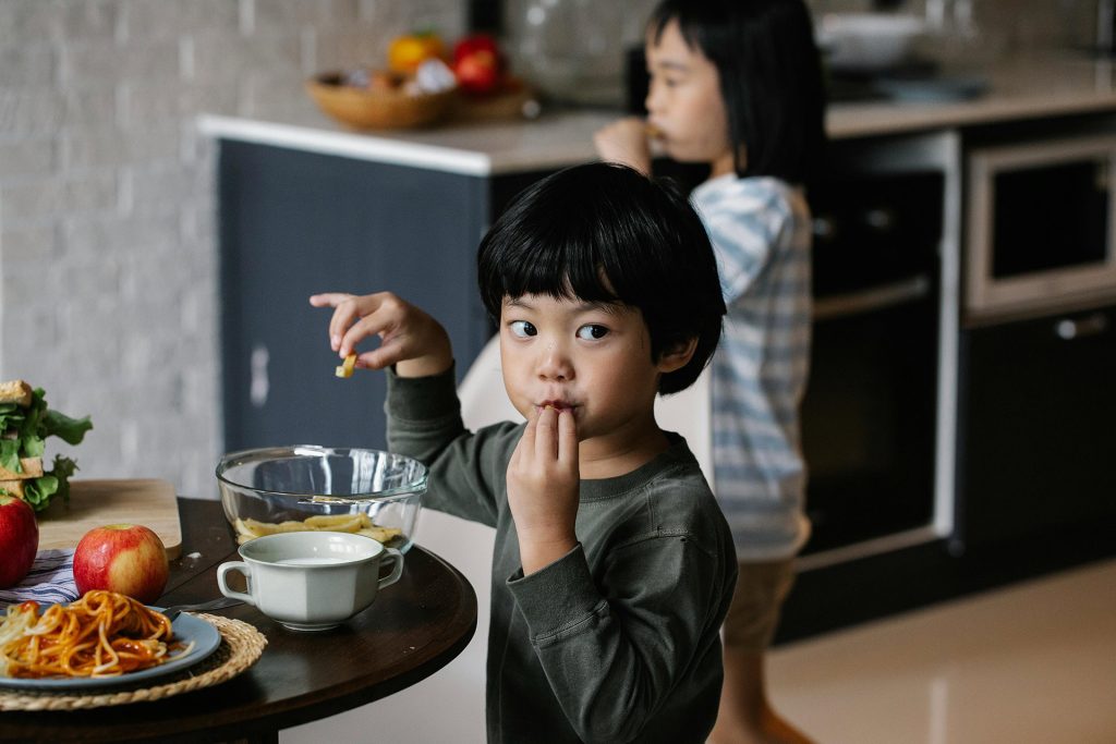 A young child eating pasta in a kitchen while an adult stands nearby, illustrating caregiver involvement in Family Based Treatment for eating disorders.