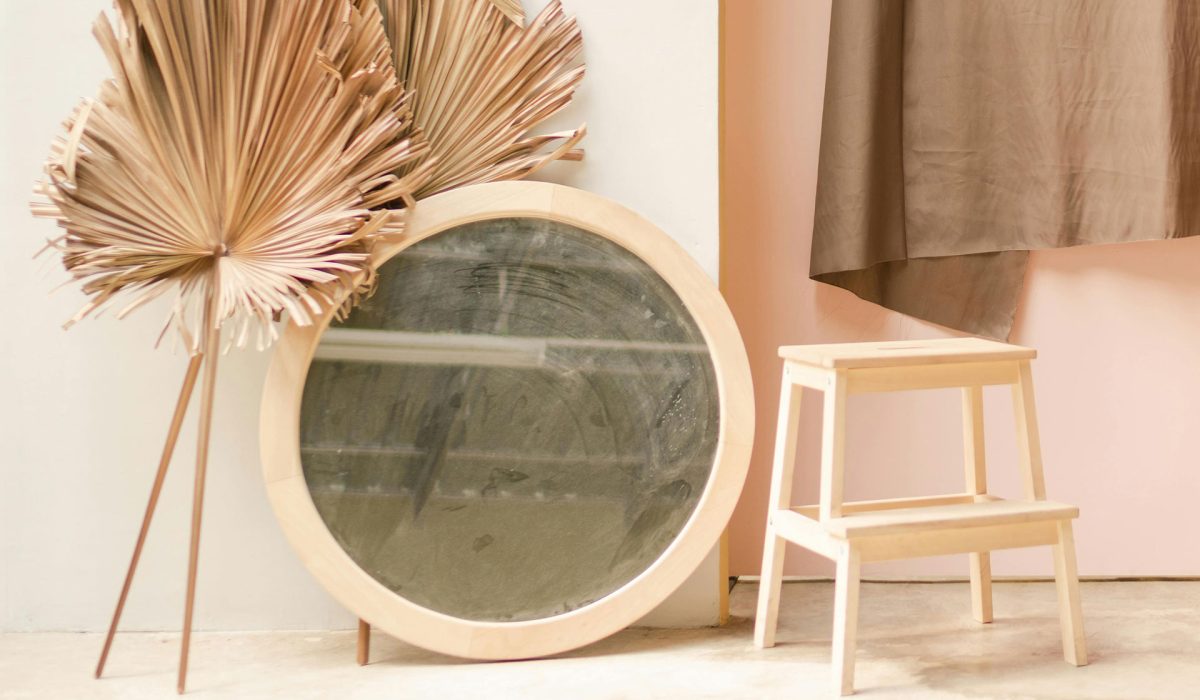 Round wooden-framed mirror leaning against a wall beside dried palm leaves and a small wooden step stool.