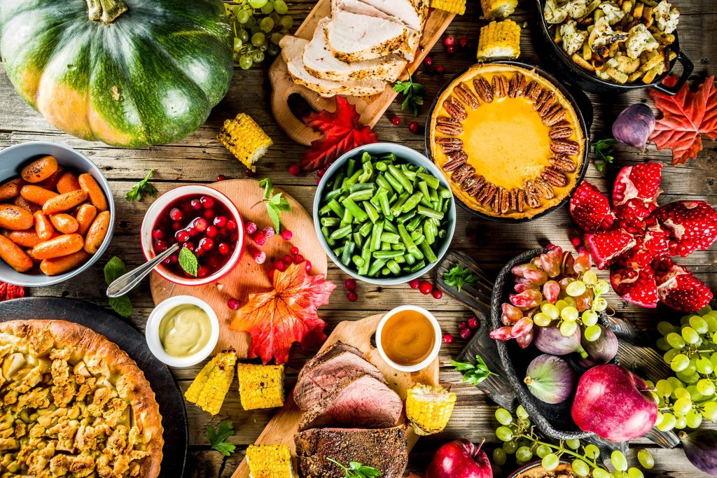 Colorful holiday meal spread featuring vegetables, roasted meats, pies, fruits, and seasonal dishes on a rustic table.
